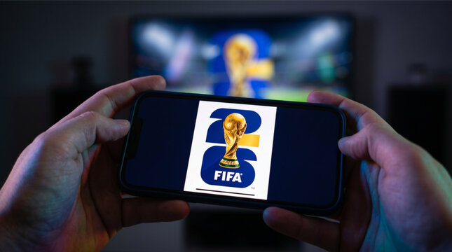 Person holding a smartphone displaying the official FIFA World Cup 2026 logo during a soccer game broadcast on TV in the background, mobile sports streaming app, football championship tournament.