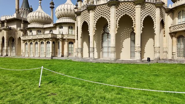 Panning shot of the historic Brighton Pavilion on a bright, sunny day, showcasing its ornate Indo-Saracenic architecture, gleaming domes, and vibrant surroundings under clear blue skies.