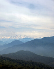 Fototapeta premium Distant snow-capped Caucasus mountain peaks behind misty blue layers of hills under a cloudy sky