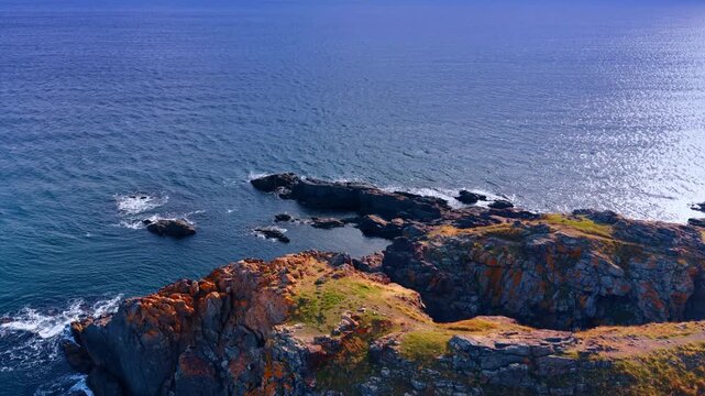 Rocky headland jutting into calm blue sea at golden hour. Elevated drone view of rugged cliff with green grass, lichen covered stones and sparkling water.