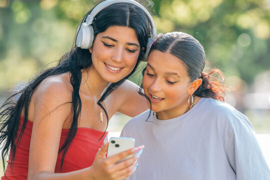 Two hispanic teenage girls share a joyful moment looking at a smartphone together in a sunny park, radiating friendship and youthful energy