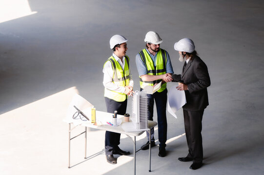 Senior businessman with architect shaking hands with young engineer at empty space floor in building construction site, real estate investors construction managers closing deal building model table.