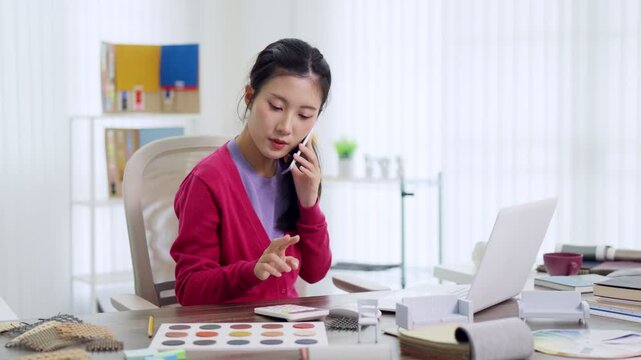 Asian woman interior designer talking phone using laptop computer reviewing furniture design material samples color swatches working home office creative