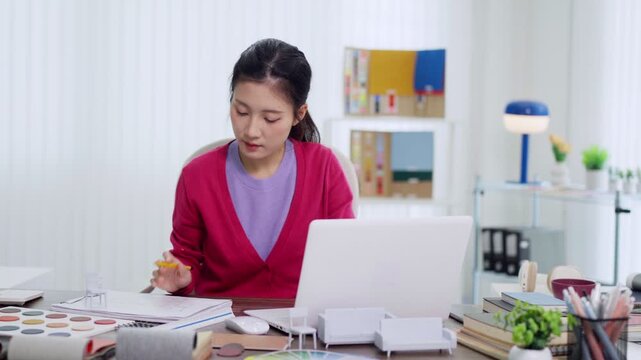 Asian female furniture designer working on creative project using laptop computer at home office desk reviewing product design plans and color samples