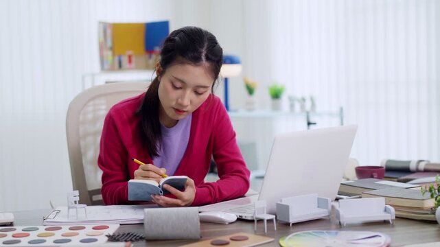 Asian designer architect taking notes in notebook reviewing furniture design plans working with architectural models at bright creative studio office desk