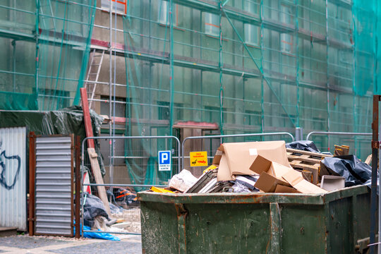A large green metal skip container filled with construction waste and cardboard boxes, located at a construction site with a building covered in scaffolding and green safety netting.