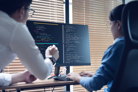 Two developers collaborate at desks with dual monitors. Code lines glow on dark screens, hinting at complex work. One points while the other types, focused on shared task