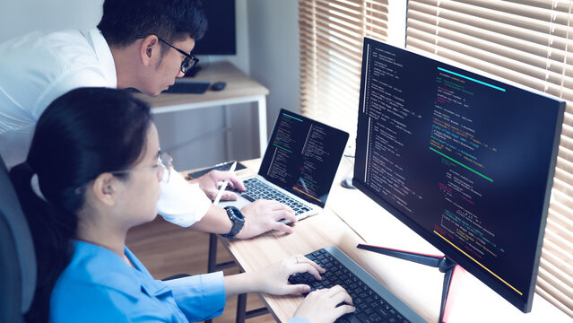 Two developers collaborate closely at their desks. One types while the other observes code on a large screen. Focused expressions suggest deep engagement with their work