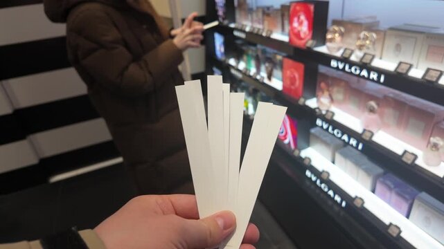 Person holds multiple white tester strips in beauty store, ready to sample various fragrances while another shopper explores products on brightly lit shelves in the background