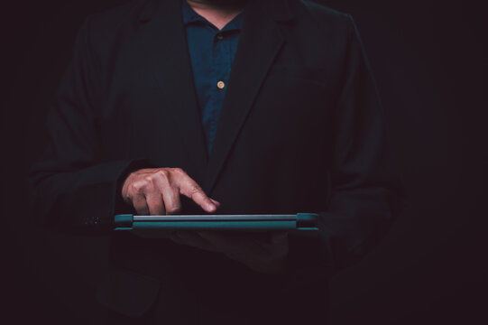 Professional businessman wearing dark suit jacket over navy shirt stands against black background tapping tablet computer screen managing digital business data inside office studio setting today.