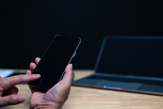 Adult male hands hold smartphone touching screen working wooden desk laptop computer dark office setting nighttime professional person browsing internet indoor workspace technology concept digital.