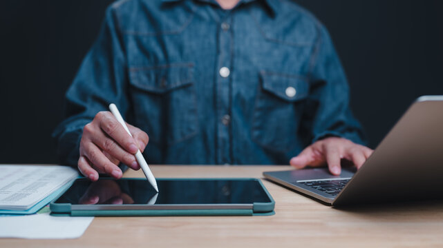 Unrecognizable adult male wearing denim shirt sits wood desk operating laptop stylus pen signing digital document tablet screen dark office night workspace setup corporate workflow technology remote.