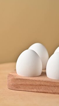 A detailed macro shot of whole white chicken eggs and one cracked eggshell revealing a fresh raw yolk inside. The wooden holder slowly rotates on a neutral beige background under soft studio lighting,