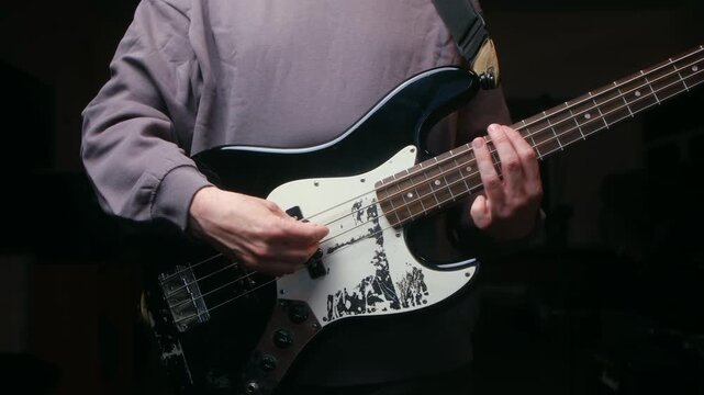 Unrecognizable musician playing four string electric bass guitar with his fingers, performing a song in a dark studio during a rehearsal. His performance shows skill and passion for music