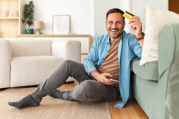 Fototapeta premium Happy smiling man holding paying credit card and mobile phone, enjoying online shopping and secure payment experience from living room at home