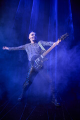 Fototapeta premium Adult male musician with electric guitar performing on dark stage with blue smoke and spotlight