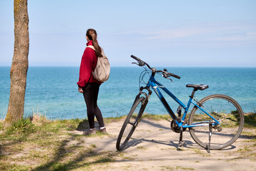 Fototapeta premium Young woman with ponytail standing by ocean shore with blue bicycle on sunny day