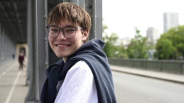 Portrait of a smiling young man wearing glasses looking directly at the camera. He stands outdoors under a bridge structure in Paris with trees in the blurred background.