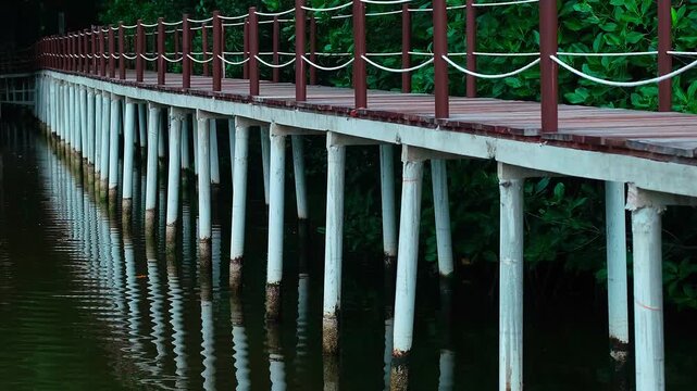 Wooden bridge spans calm, green water. White pillars support the walkway gently. Red railings and rope accents add color. Reflections ripple softly in the water