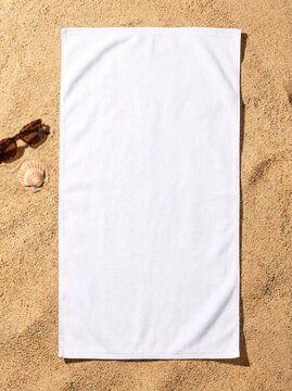 A blank white beach towel mockup flat lay on sand with sunglasses and a seashell for custom summer merchandise design