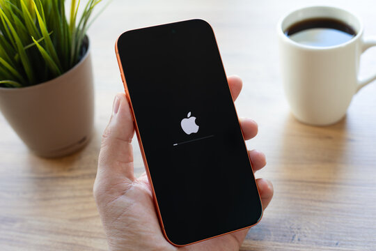 Man hands holding Apple iPhone 17 Pro Max Cosmic Orange showing the Apple logo on a black boot screen, on desk with coffee in the background