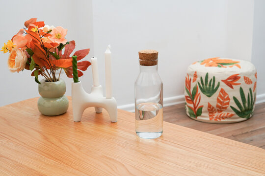 A stylish still life of modern home decor on a light wood table. The scene features a vase of orange flowers, a white candelabra, and a glass water bottle. A patterned pouf sits in the background.