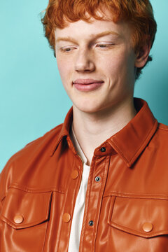 Young redhead man with smile and closed eyes wearing brown leather jacket and white shirt standing against blue background in studio portrait for fashion and lifestyle.
