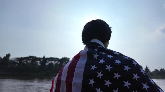 Cinematic slow motion tracking shot zooming out from a man wearing the star-spangled banner on his shoulders, revealing a wide scenic view of a quiet river and local landscape under bright daylight.
