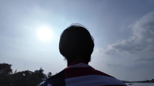 Close up tracking shot focusing on the back of a man's head and hair as he wears the United States flag, looking upward against a bright sun flare and a vast blue sky near a lakeside.
