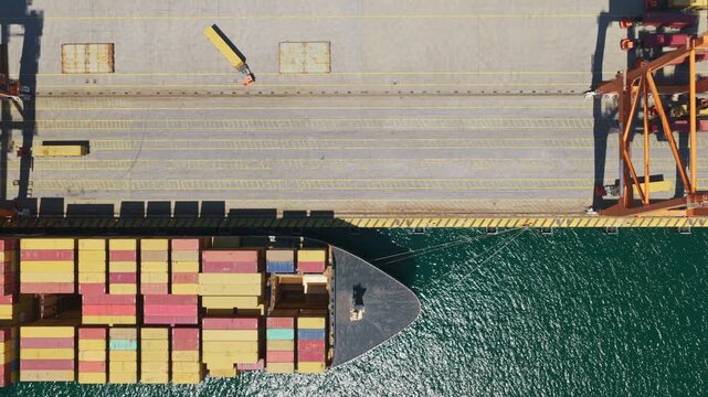 Industrial port scene featuring an aerial top-down view of a massive container cargo ship slowly moving along the pier, showcasing the scale of global trade and maritime logistics operations