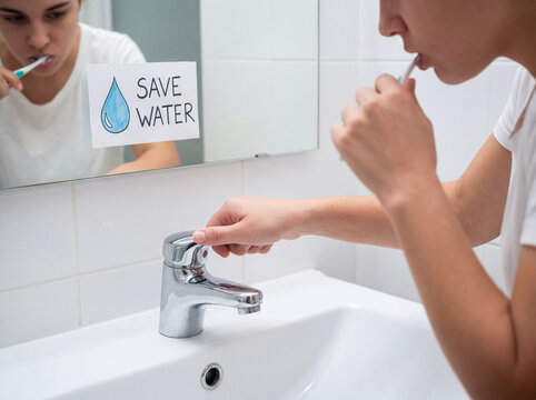 Woman turning off a water faucet while brushing her teeth, actively conserving water with a reminder in the mirror