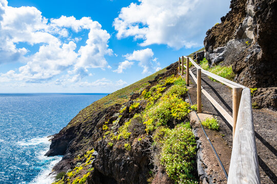 Dirt hiking path with a wooden railing winds along a high volcanic cliff with sweeping ocean views at Nogales beach, La Palma, Canary Islands, Spain