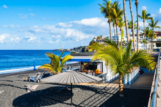 Thatched umbrella and palm trees shade a lively beachside kiosk on a black sand shore under a blue sky in Puerto Naos town, La Palma, Canary Islands, Spain
