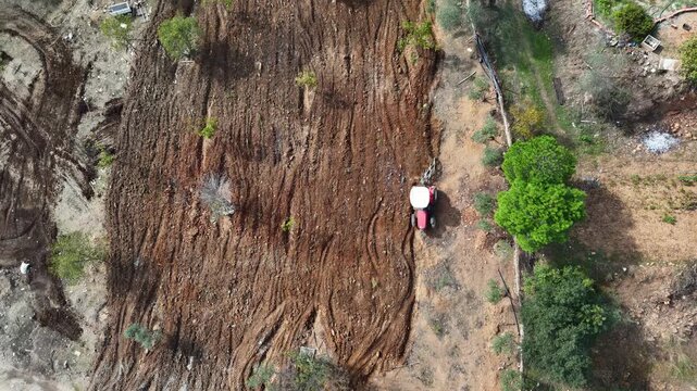Aerial drone shot of a red tractor working on tilled brown soil in rural Turkey, surrounded by green trees and natural landscape.