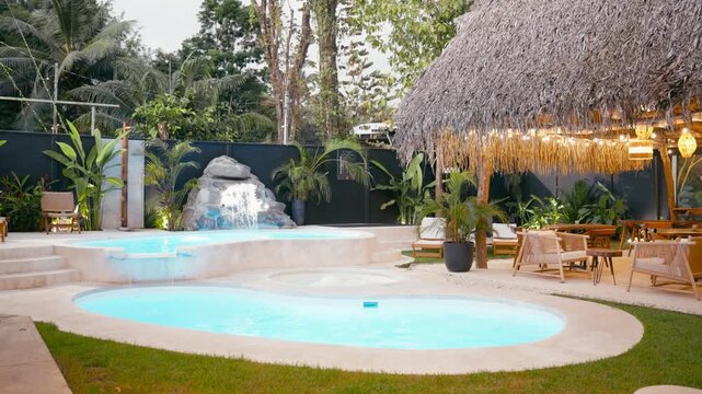 Beautiful view of a luxury hotel swimming pool area with a small waterfall, jacuzzi, and a tiki style bar surrounded by tropical nature