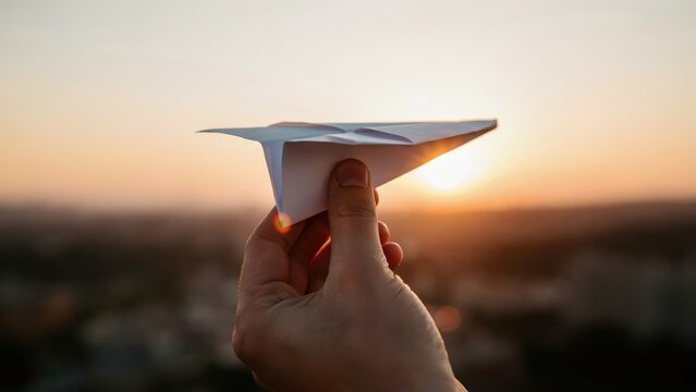 Hand holding paper airplane against a golden sunset sky, symbolizing hope and freedom.
