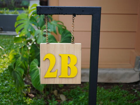 House number sign 2B hanging on metal chains featuring yellow letters attached to a wooden square board outdoors garden background