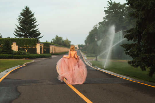 A full length back view of a woman in a flowing pink tulle dress running along an empty asphalt road.
