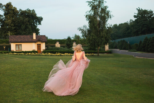 A young woman in a gorgeous fluffy pink dress running away across a wide green lawn towards a house.
