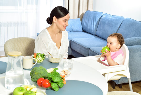 Mother feeding baby with a spoon. Spoon-feed. Happy young mother with a baby in the kitchen interior. Fresh vegetables and fruits.