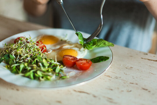 A close up of a person using a fork to pick up fresh green spinach leaves from a plate with fried eggs and microgreens.