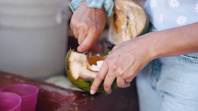 Close up of female hands scraping the sweet pulp from a green coconut with a spoon. A young girl's head appears, ready to enjoy the snack