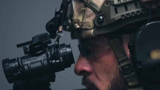 Male soldier in tactical gear adjusts night vision scope on rifle, focusing intently with a serious expression in a dimly lit indoor environment