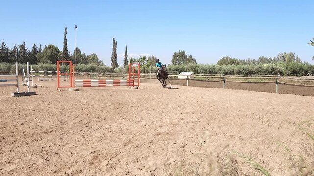 ​A dynamic, ground-level medium shot of a young equestrian rider, a teenager, cantering a powerful dark bay horse through a turn in a dirt training arena. 