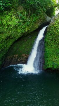 Aerial view of a hidden waterfall deep within the lush tropical rainforest of Mount Daun, Indonesia. Surrounded by dense green jungle vegetation, the pristine cascade flows into a dark natural pool