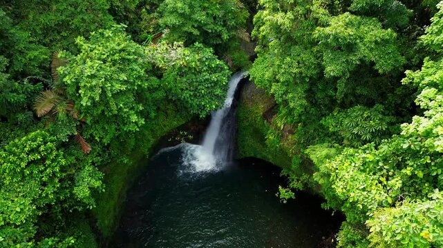 Aerial view of a hidden waterfall deep within the lush tropical rainforest of Mount Daun, Indonesia. Surrounded by dense green jungle vegetation, the pristine cascade flows into a dark natural pool