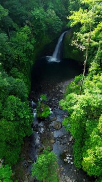 Aerial view of a hidden waterfall deep within the lush tropical rainforest of Mount Daun, Indonesia. Surrounded by dense green jungle vegetation, the pristine cascade flows into a dark natural pool