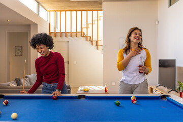 Obraz premium Diverse female friends laughing and playing pool at home with cue stick, holding beverage