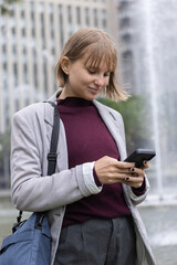 Obraz premium Adult woman standing in plaza by fountain holding smartphone wearing blazer and blue crossbody bag