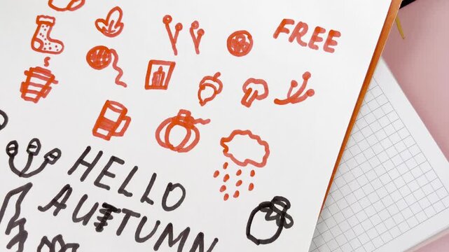 Creative person doodling autumn themed icons and writing hello autumn in a notebook. Seasonal planning concept featuring pumpkins, acorns, rainy clouds, and warm drinks